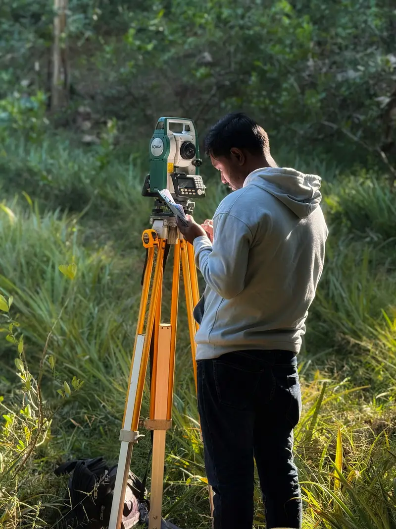 A surveyor reading a total station set up on a tripod in tall grass